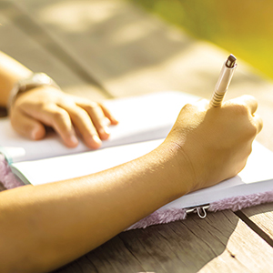 A person writes in a notebook with a pen while sitting at a wooden table outdoors in bright sunlight.