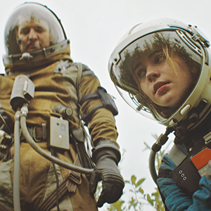 Two people in bulky space suits with helmets stand outdoors, surrounded by greenery. One person looks down thoughtfully while the other gazes into the distance. The sky is overcast.