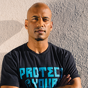 A bald man with medium skin tone stands against a textured wall, arms crossed, wearing a black T-shirt that says PROTECT YOUR in bold blue letters. He looks confidently at the camera.