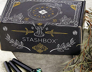 A decorative black box labeled STASHBOX sits on a textured surface beside three black vape pens with colorful mouthpieces and dried foliage. The box features gold and white mystical designs.