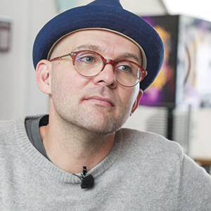 A person wearing a blue hat, brown-rimmed glasses, and a light gray sweater looks slightly to the side with a thoughtful expression. The background is softly blurred with colorful abstract shapes.