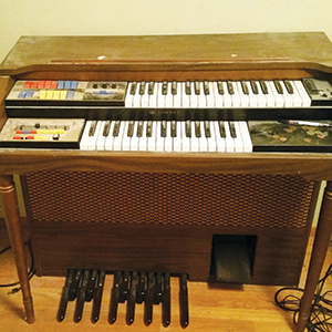 A vintage electric organ with two keyboard rows, colorful buttons, foot pedals, and a wooden cabinet standing on a hardwood floor.