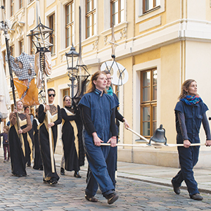 A group of people parade down a cobblestone street, some dressed in dark gowns and others in blue coveralls, carrying large lanterns and props. The backdrop is a yellow, historic building with tall windows.