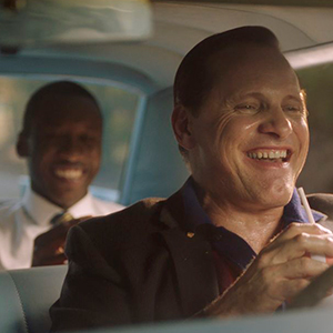 Two men are inside a car; one is driving and smiling, while the other sits in the backseat, also smiling. Sunlight shines through the windows, creating a warm atmosphere.