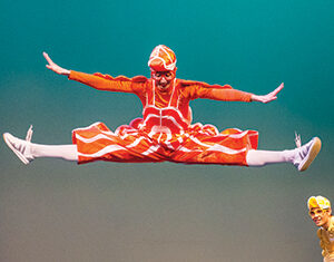 A performer in a bright orange costume and hat does a high mid-air split jump on stage, while another performer in yellow watches from the side against a plain green background.