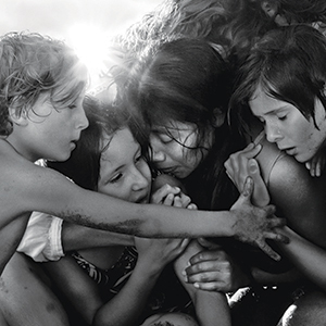 A black-and-white photo of four children and a woman huddled closely together, embracing each other with worried, emotional expressions. Sunlight shines in the background, highlighting the groups intense moment.