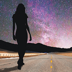 Silhouette of a person walking down an empty road toward distant mountains, with a vibrant, star-filled night sky overhead.