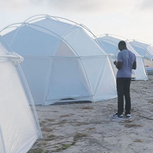 A person stands outside white dome-shaped tents set up on rough ground under a cloudy sky.