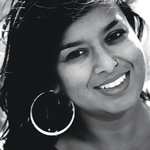 A woman with long dark hair, wearing large hoop earrings and a nose ring, smiles at the camera. The image is in black and white.