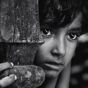 A young child with expressive eyes gazes pensively while partially hiding behind a wooden post, gripping it with one hand. The image is in black and white, highlighting the child’s thoughtful expression.