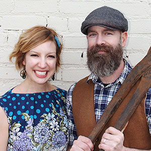 A smiling woman in a blue floral dress stands next to a bearded man wearing a flat cap, plaid shirt, and brown vest, holding wooden skis, in front of a white brick wall.