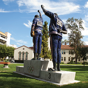 A sculpture of two athletes on Olympic podiums, each raising a clenched fist. They stand on first and third place blocks, wearing numbered uniforms, with trees and campus buildings in the background.