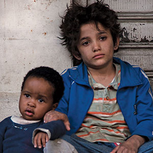 Two young children sit on the ground against a wall. The older child, with messy hair and a blue jacket, looks ahead with a serious expression. The younger child, sitting close, rests their arm on the older child’s leg.