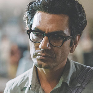 A man with dark hair and glasses looks serious. He is wearing a light grey shirt and standing indoors with blurred people and lights in the background.