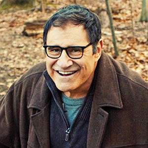 A man with dark hair and glasses, smiling outdoors in a wooded area during autumn. He is wearing a dark coat over a sweater, with dry leaves scattered on the ground behind him.