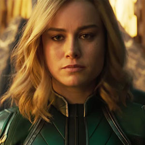 A woman with shoulder-length blonde hair wearing a green and gold superhero suit stands facing forward, looking serious and determined, with a blurred background.