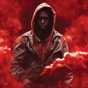 A person wearing a hooded jacket stands in a cloud of red smoke, holding a red flare, with an intense expression on their face. The background is filled with swirling red smoke.