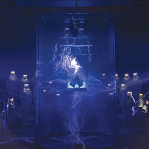 A person sits in a glass chamber surrounded by blue electricity and lightning effects, while people in hazmat suits watch from the background in a dark, dramatic setting.