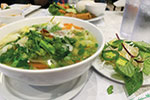 A bowl of Vietnamese pho soup with herbs and vegetables on a plate beside it, served on a white table with other dishes and a glass of water in the background.