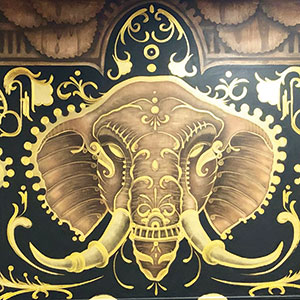 Ornate painting of an elephants head in gold and brown tones, featuring decorative patterns and flourishes on a dark background. The artwork is symmetrical and highly stylized.