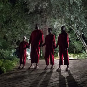 Four people wearing matching red outfits stand hand-in-hand on a brick path at night, surrounded by trees and illuminated by soft, dramatic lighting. Their faces are in shadow, creating a mysterious atmosphere.
