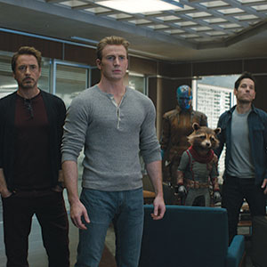 Five Marvel superheroes, including a man in a gray shirt, a man in glasses, a blue-skinned character, a humanoid raccoon, and another man, stand together indoors, looking intently ahead.