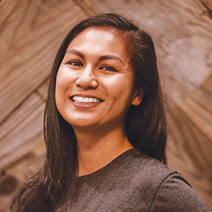 A woman with long dark hair smiling, wearing a dark shirt, standing in front of a wooden textured background.