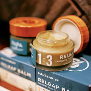 An open jar of Papa & Barkley Releaf Balm sits on top of its blue and white packaging, with another jar and lid blurred in the background. The balm is creamy yellow in color.