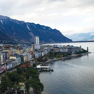 A lakeside town with colorful buildings, a high-rise, and tree-lined streets sits beside calm water, with tall mountains in the background under a partly cloudy sky.