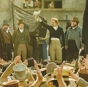 Four men in 19th-century attire stand on a platform before a cheering crowd; one man raises his hat in the air while others look on, and a dark banner is visible in the background.