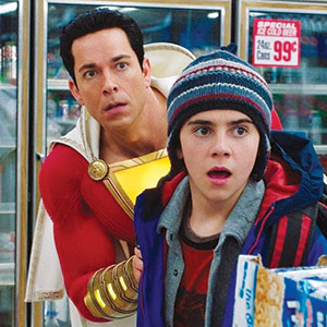 A man in a superhero costume and a surprised boy in a striped beanie stand in a grocery store, looking ahead with shocked expressions. Frozen food and a price sign are visible in the background.