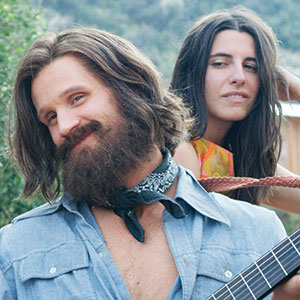 A bearded man with long hair, wearing a blue denim shirt and bandana, smiles while holding a guitar. A woman with long brown hair stands behind him, looking to the side, with greenery in the background.