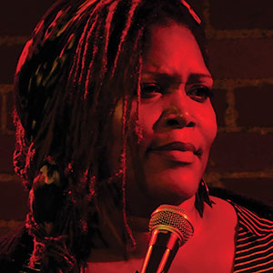 A person with long, braided hair stands in front of a brick wall, holding a microphone and looking thoughtful, bathed in red stage lighting.