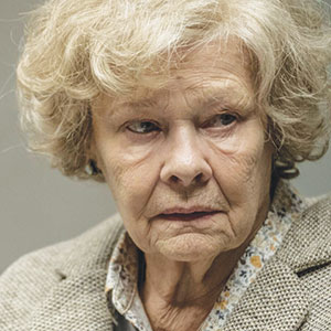 An elderly woman with curly, light gray hair and a serious expression is wearing a light-colored blazer over a patterned blouse, looking slightly to the side.
