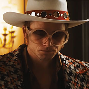 A man wearing a large white cowboy hat, oversized round glasses, and a patterned jacket stands indoors with a serious expression; warm lighting is visible in the background.