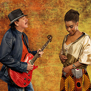 A man in a hat plays a red electric guitar while standing next to a woman with braided hair, wearing a patterned skirt and looking down. The background is a textured orange and yellow.