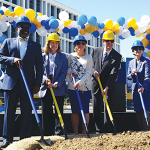 Five people in suits and yellow or blue construction helmets holding shovels at a groundbreaking ceremony, with colorful blue, white, and yellow balloons in the background.