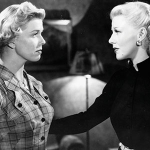Two women stand facing each other indoors; one wears a plaid dress, the other a dark top with earrings, and gently places a hand on the others arm. The scene is in black and white.