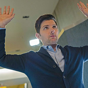 A man with short dark hair, wearing a blue cardigan and light shirt, stands indoors with both hands raised above his head, looking slightly upwards with a serious expression.