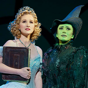 Two women stand side by side in theatrical costumes. One, with blonde curls and a blue dress, holds a book. The other, with green skin and a black pointed hat and dress, looks ahead. They appear to be on stage in a musical.