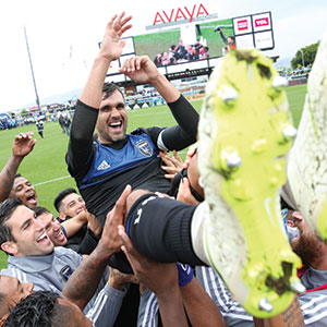 A group of soccer players lift and celebrate a teammate, who is smiling and raising his arm, on a soccer field with a stadium scoreboard in the background.