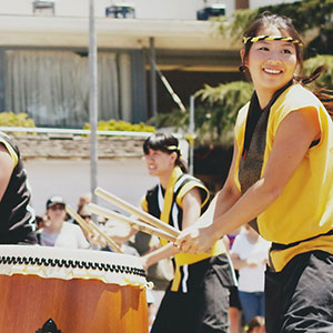 A group of people in yellow and black outfits joyfully play large traditional drums outdoors, with a crowd watching in the background.
