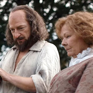 A man with long hair and a beard, dressed in old-fashioned clothing, stands next to an older woman with curly hair in a period dress. They appear to be outdoors, with trees blurred in the background.