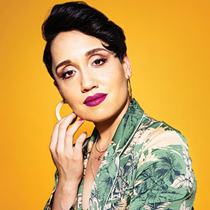 A person with short dark hair, bold pink lipstick, and hoop earrings touches their face. They wear a green floral blazer and pose against a bright yellow background.