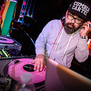 A DJ wearing glasses, a gray hoodie, and a cap that says Cumbia Cartel mixes music on pink vinyl turntables in a colorful, dimly lit setting.