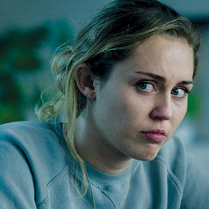 A young woman with light brown hair pulled back looks to the side with a sad, teary expression. She is wearing a grey sweatshirt and appears upset, with a slightly red eye and a somber background.