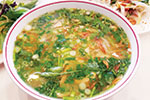 A bowl of Vietnamese pho soup filled with noodles, fresh herbs, green onions, and slices of meat in a clear, flavorful broth. A plate of garnishes is visible in the background.