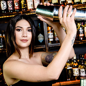 A woman with shoulder-length dark hair shakes a cocktail shaker behind a bar lined with bottles of liquor. She has tattoos on her arm and is wearing a sleeveless top, looking confidently at the camera.