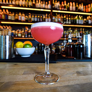 A pink cocktail with a foamy top served in a coupe glass sits on a bar counter; liquor bottles and a bowl of citrus fruits are visible in the background.