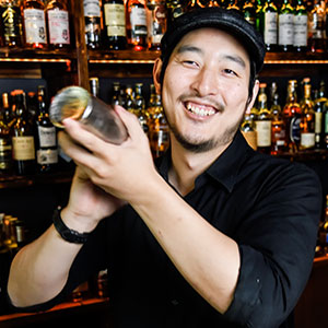 A smiling bartender wearing a black shirt and cap shakes a cocktail shaker behind a bar lined with various bottles of liquor.